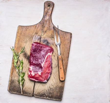 raw ribeye steak on a cutting board with a vintage meat fork and a sprig of rosemary on a white wooden rustic backgroundの写真素材