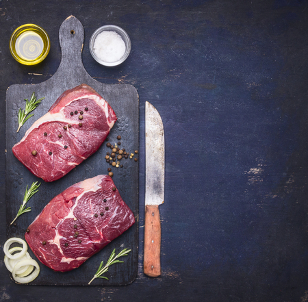 Raw beef steak with rosemary, butter and meat fork on dark rustic cutting board, top view with space for textの写真素材