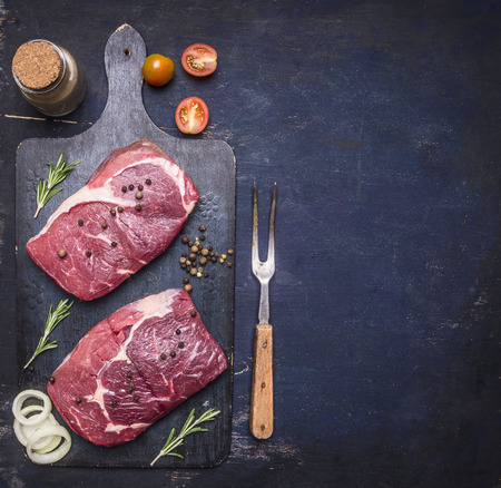 two raw ribeye steak on vintage cutting board with rosemary, herbs, pepper and meat fork, Border, place for text on a rustic wooden backgroundの写真素材