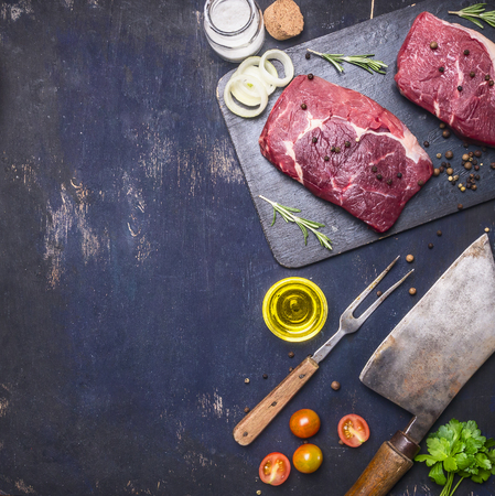 concept of cooking meat, raw ribeye steak on a cutting board with rosemary, spices, oil and vegetables, with a meat cleaver vintage, Border, place for text on a rustic wooden backgroundの写真素材