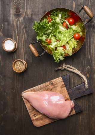 Fresh salad with cherry tomatoes, spices and herbs, in a copper pot, with raw chicken breast on a cutting board, sports dinner on wooden rustic background top viewの写真素材