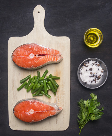 preparing raw salmon steak with herbs, salt, pepper and other seasonings, two steaks  lie on the cutting  board, on a black background a rustic close upの写真素材