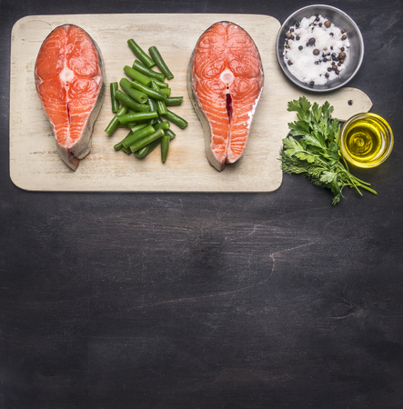 preparing raw salmon steak with herbs, salt, pepper and other seasonings, two steaks  lie on the cutting  board, on a black background a rusticの写真素材