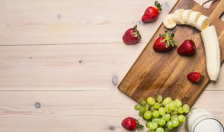 Cooking homemade smoothies made from strawberries and bananas with grapes, ingredients are laid out on a cutting board on a white wooden rustic background, border, space for textの写真素材
