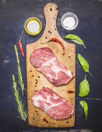 Two raw appetizing pork steaks on a vintage cutting board with meat knife, garlic, salt and spicy pepper, butter and rosemary and basil on a rustic wooden backgroundの写真素材