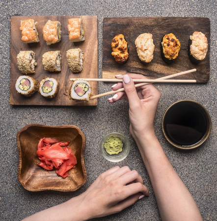 women's hands take sushi with chopsticks, ginger, wasabi, soy sauce, various rolls are laid out on a rustic granite backgroundの写真素材