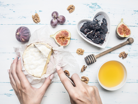women's hands cut off a piece of camembert cheese on white packing paper, with jam, walnuts and honey, freshly cut figs, on a rustic wooden backgroundの写真素材