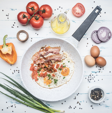 homemade scrambled eggs with bacon, tomatoes, green onions, bell pepper and seasonings on a white frying pan, around lie the ingredients top viewの写真素材