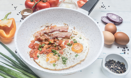 homemade scrambled eggs with bacon, tomatoes, green onions, bell pepper and seasonings on a white frying pan, around lie the ingredientsの写真素材