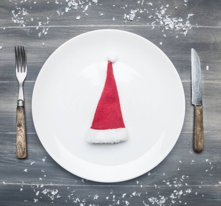 white plate with a Christmas hat on a wooden rustic table, with vintage cutlery, and snow, top view, space for textの写真素材