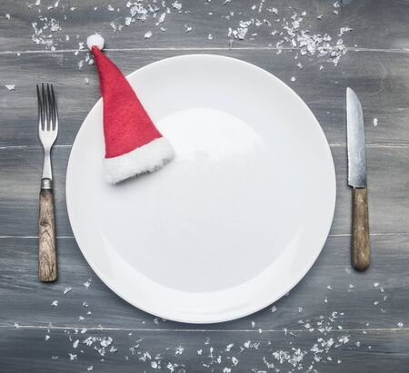 white plate with a Christmas hat on a wooden rustic table, with vintage cutlery, and snow, top view, space for textの写真素材