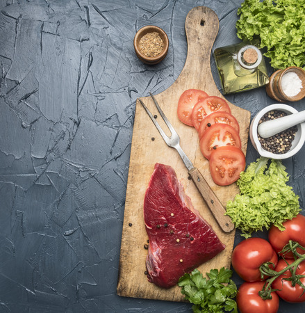 appetizing raw beef steak with tomatoes and fork for meat on a vintage cutting board, surrounded by ingredients, spices and spices, on rustic dark background, space for textの写真素材