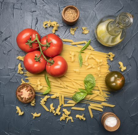 different types of pasta, laid out with various ingredients, tomatoes, herbs, oil in a decanter and seasonings on a dark rustic background, healthy vegetarian foodの写真素材