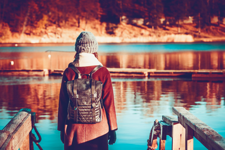 Girl traveler in autumn clothes, long hair and a backpack behind her shoulders, stands on the lakeside by the lake, against the background of yellow trees and waterの写真素材