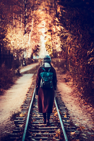 Girl in a burgundy coat and a gray hat, with a leather backpack on her back, walking along an abandoned railway, against the backdrop of an autumn landscape, yellow leaves and trees, blureの写真素材