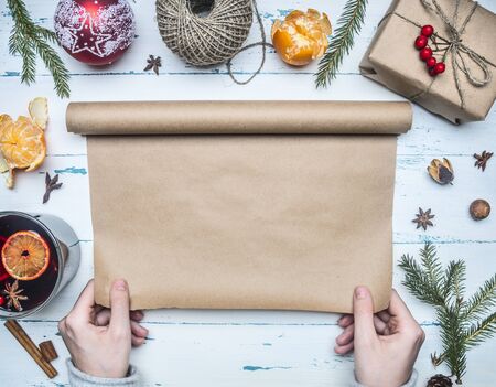 girl in sweater unwinds a roll of wrapping paper for wrap-up for a gift in it for a new year or Christmas, a glitwin cup, twigs of a tree, mandarins, a string, a wooden rustic background, a place for textの写真素材