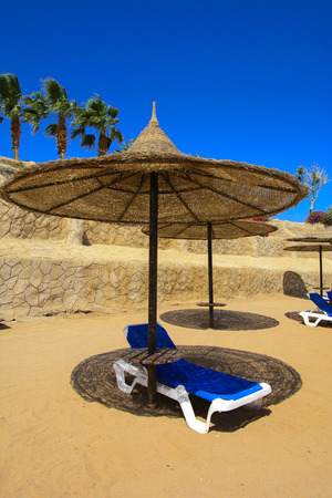 A row of straw umbrellas to protect against overheating and sunbeds on a sandy beach against a blue sky and blue sea. The concept of summer holidays, tourism, travel.の写真素材