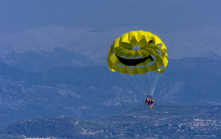 A skydiver flies with a colorful parachute tied to a speedboat on the sea against the backdrop of mountains with a blue sky. The concept of summer holidays, vacation, tourism.のeditorial素材