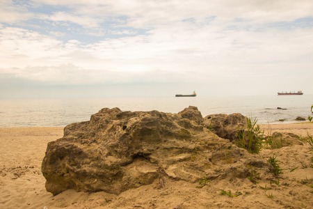 Big rock on the sunny beach with blue turquoise sea and cloudy sky and cargo shipsの写真素材