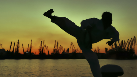 martial artist training alone on the sea pier, practicing his moves on sunset and port cranes background.の写真素材