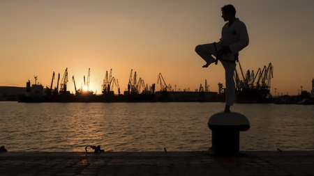 martial artist training alone on the sea pier, practicing his moves on sunset and port cranes background.の写真素材