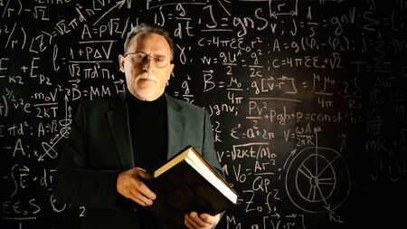 Medium shot of male university professor with book in hands. Math formulas on blackboard. Knowledge and On-line education concept.の写真素材