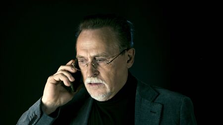Mature gentleman talking on a smartphone. Medium shot portrait elderly elegant man with phone on dark background. Handsome gray haired old businessman in glasses.の写真素材