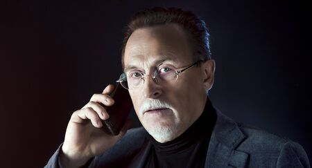 Mature gentleman talking on a smartphone. Medium shot portrait elderly elegant man with phone on dark background. Handsome gray haired old businessman in glasses.の写真素材