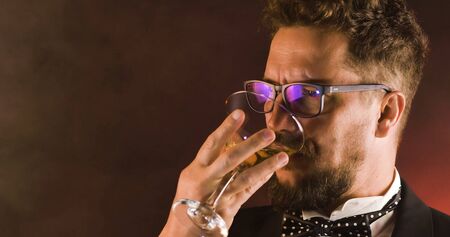 Handsome man in suit drinking champagne on black background. Portrait of elegance gentleman with glasses. Confident adult man in suit holding glass of champagne in studio. Luxury party.の写真素材