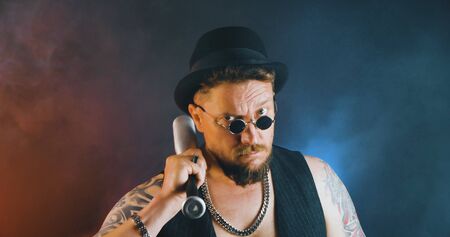 Portrait of athletic guy with a tattoo poses with a baseball-bat in dark studio. Cool mafia gangster in sunglasses and cylinder hat. Bearded bouncer with baseball-bat and sunglasses.の写真素材