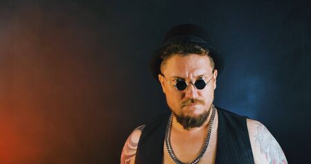 Portrait of athletic guy with a tattoo poses with a baseball-bat in dark studio. Cool mafia gangster in sunglasses and cylinder hat. Bearded bouncer with baseball-bat and sunglasses.の写真素材
