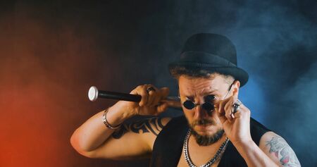 Portrait of athletic guy with a tattoo poses with a baseball-bat in dark studio. Cool mafia gangster in sunglasses and cylinder hat. Bearded bouncer with baseball-bat and sunglasses.の写真素材