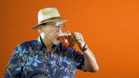 Mature joyful man in summer hawaiian shirts and straw hat with a mug of beer on a orange background.