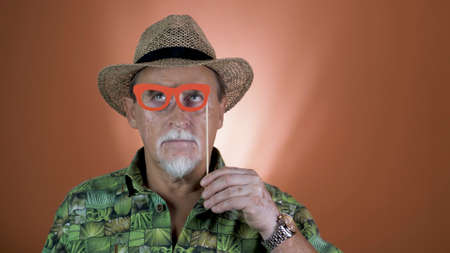 Studio portrait of the joyful elderly tourist in a straw hat and hawaiian shirt on colored backgrounds. Peoples emotion. Happy and funny celebrating people.の写真素材