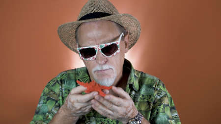 Portrait of a joyful elderly tourist in a straw hat and hawaiian shirt holding a starfish on a...