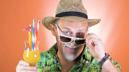 Portrait of a joyful elderly tourist in a straw hat and hawaiian shirt holding a tropical orange juice use on a colored background.の写真素材