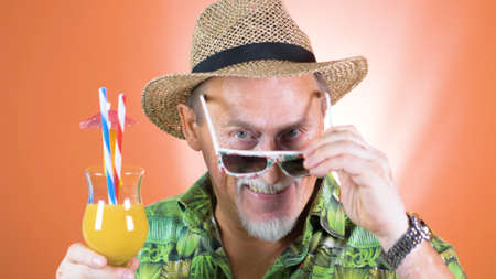 Portrait of a joyful elderly tourist in a straw hat and hawaiian shirt holding a tropical orange juice use on a colored background.の写真素材