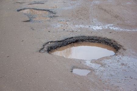 The bad asphalted road with a big pothole filled with water. Dangerous destroyed roadbed. Mirror reflections in the water.