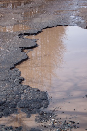 Dangerous destroyed roadbed. The bad asphalted road with a big pothole filled with water. Mirror reflection trees in the water.の写真素材