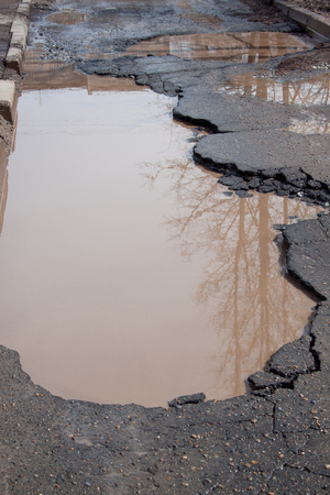 The bad asphalted road. Big pothole filled with water. Dangerous destroyed roadbed. Mirror reflection trees in the water.の写真素材