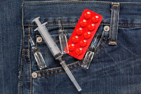Injection syringe with ampullas and red packaging with pills are lying in side pocket of jeans. Medical health or drugs addiction concept.の写真素材