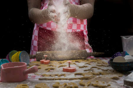 Girl claps hands and spraying flour over dough on dark background. Professional girl baker in apron is preparing cookiesの写真素材
