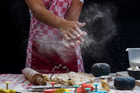 Girl claps hands and spraying flour over dough on dark background. Professional girl baker in apron...