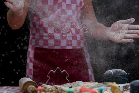 Girl claps hands and spraying flour over dough on dark background. Professional girl baker in apron is preparing cookiesの写真素材
