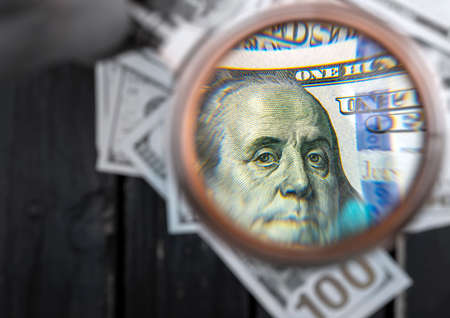 One hundred dollar bill being studied through a magnifying glass on a old black wooden table. Selective focusの写真素材