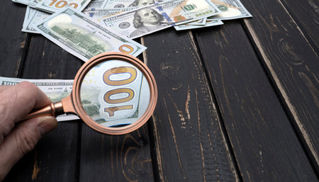 One hundred dollar bill being studied through a magnifying glass on a old black wooden table....