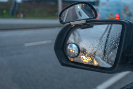 Raindrops on car window with blurred side mirror and city lights, abstract rainy day transport background for weather, traffic safety and insurance themes.の写真素材