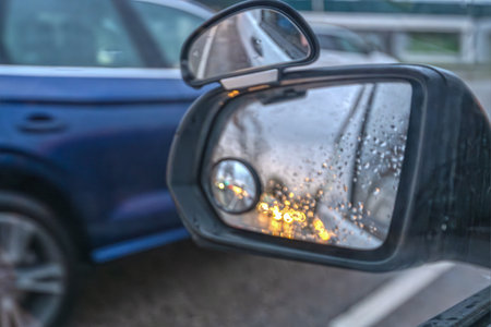 Raindrops on car window with blurred side mirror and city lights, abstract rainy day transport background for weather, traffic safety and insurance themes.の写真素材