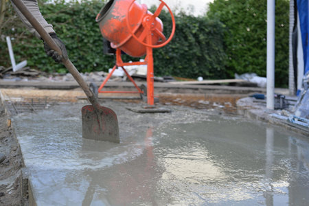 Close-up of a builder in rubber boots pouring and spreading wet concrete into wooden formwork for a slab. Construction site concreting and foundation workの写真素材
