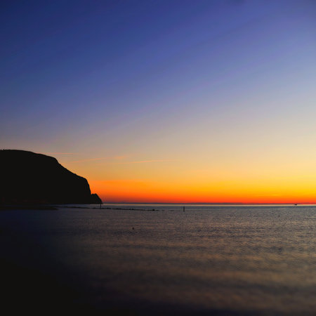 This awe-inspiring photo features the stunning coastline at sunrise, with a magnificent mountain silhouette in the background. The brilliant blue and orange hues of the sky create a captivating contrast against the calm, deep blue sea. The texture of the water, with its gentle waves and reflections of light, adds a sense of movement and depth to the image. The mountain's silhouette, with its craggy peaks and rugged terrain, adds a sense of grandeur and majesty to the scene. The photo captures a moment of stillness and beauty, inviting the viewer to contemplate the wonders of nature and the power of the rising sun. The location, unknown but assumed to be coastal, adds an air of mystery and adventure to the image, evoking a desire to explore and discover. Overall, the photo is a testament to the beauty and power of nature and the human spirit's capacity for awe and wonder.の写真素材